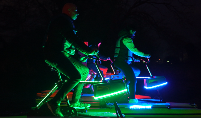 Two individuals on illuminated bicycles in a dark setting.