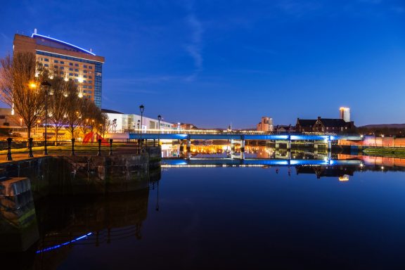 Cityscape at dusk with a river reflecting buildings and lights.